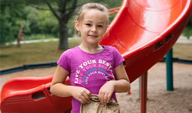 A smiling girl stands in front of a red slide at a park, holding a paper bag.
