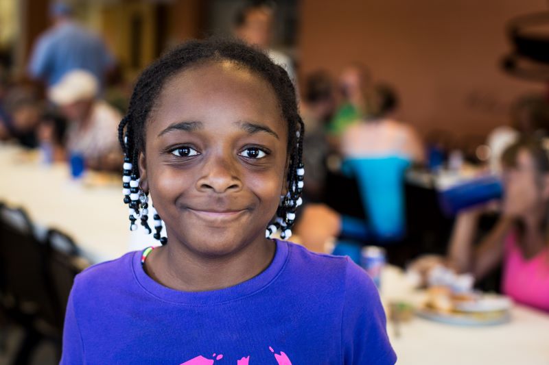 A smiling girl stands in front of a red slide at a park, holding a paper bag.